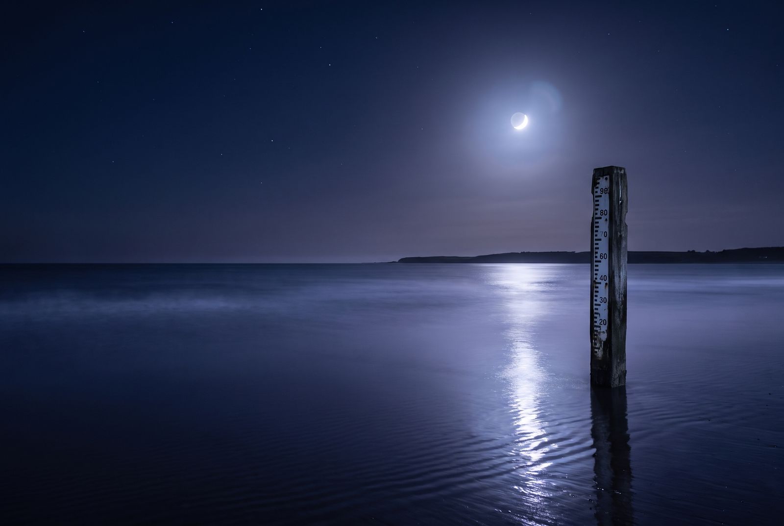 A wooden tide gauge standing in a calm night sea under a crescent moon